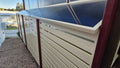 A solar panel kit mounted on a cream-colored aluminum slatted fence with dark red posts, installed outdoors next to a gravel path.