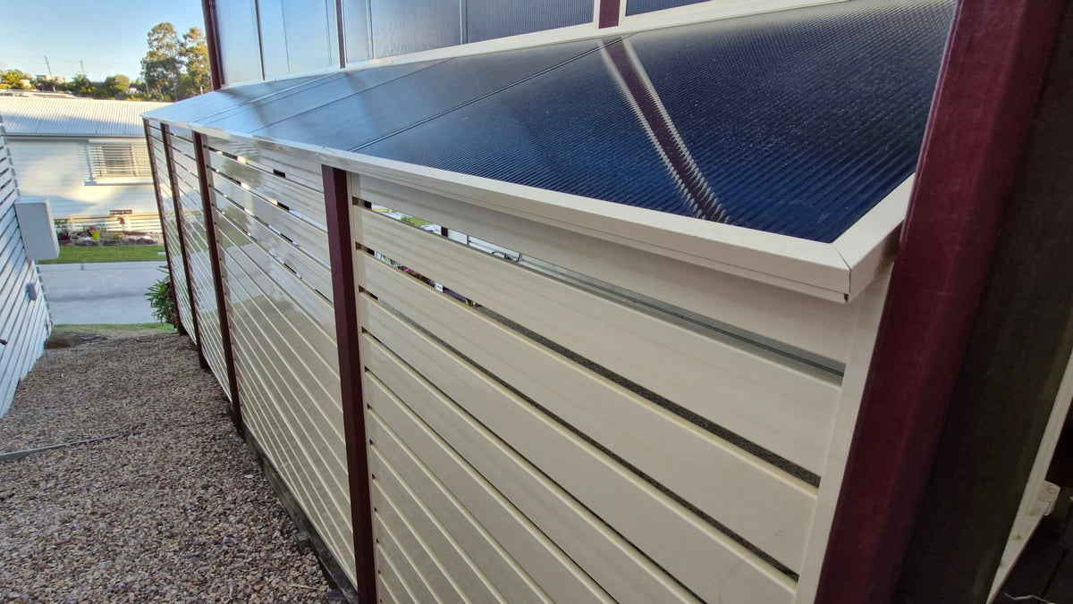 A solar panel kit mounted on a cream-colored aluminum slatted fence with dark red posts, installed outdoors next to a gravel path.