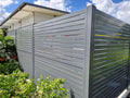 A modern gray metal slat fence installed along the side of a house, with green foliage in the foreground and a blue sky with clouds in the background.